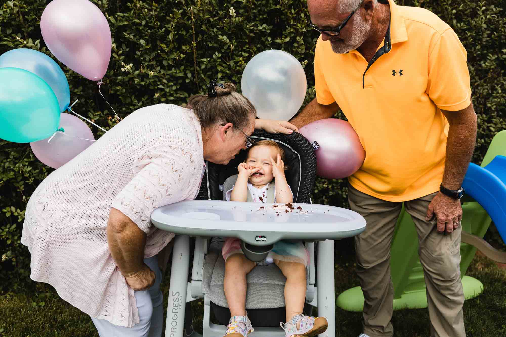 little girl sits in her high chair eating her smash cake at her first birthday party with family and balloons around her